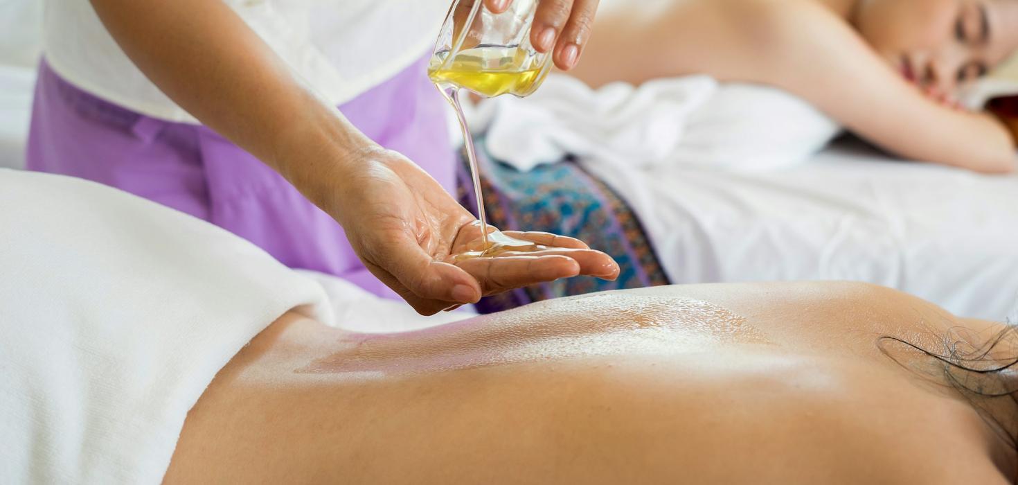 Eine Therapeutin schüttet sich öl auf eine Hand, um eine Frau zu massieren. Im Hintergrund liegt eine weitere Frau auf einer Massageliege.